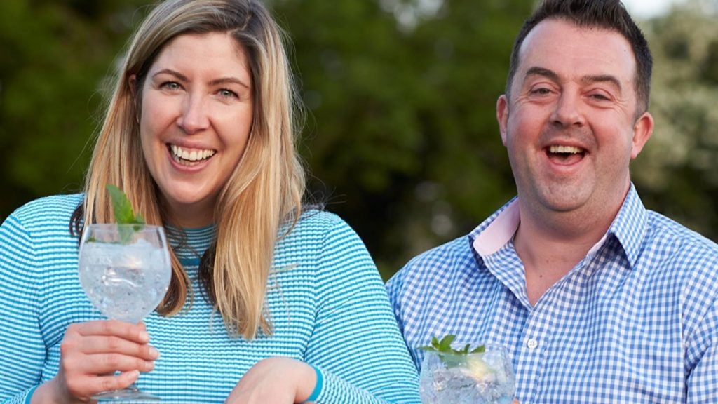 woman with blonde hair and man with black hair in blue coloured clothing laughing at camera holding glasses of gin.